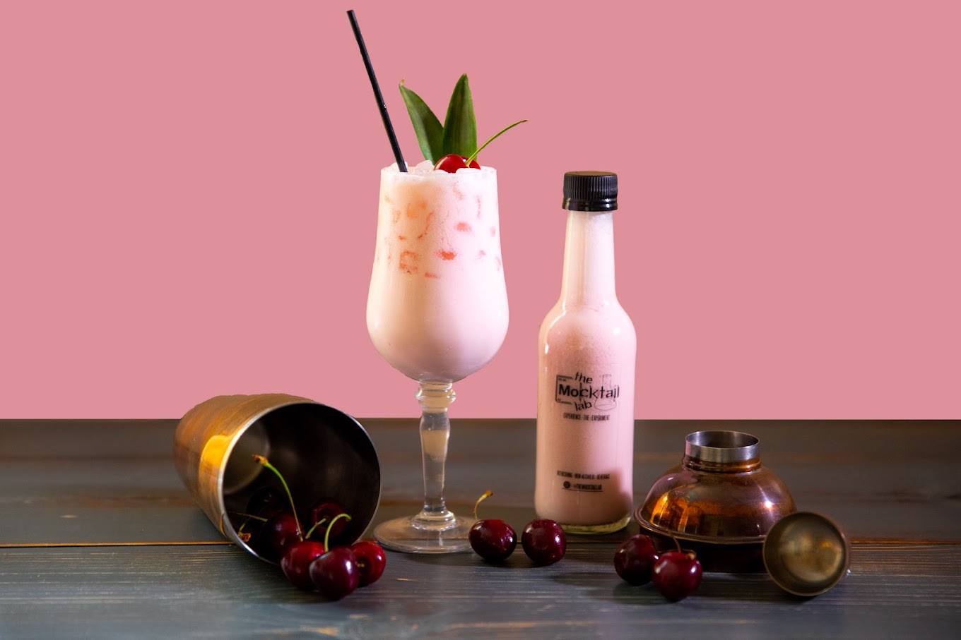 Pink signature mocktail garnished with cherry and pineapple leaf, served beside a bottled batch
